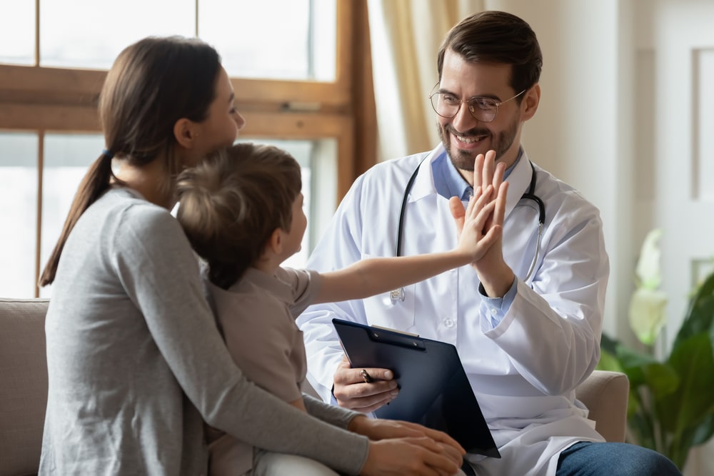 Smiling male pediatrician giving high five to young boy during clinic visit with mother, highlighting positive child healthcare experience.