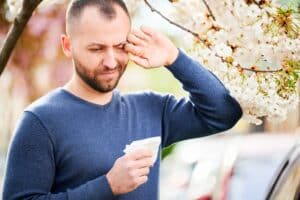 Man sneezing and rubbing itchy eyes near blooming tree, showing symptoms of seasonal spring allergies