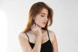 Young woman with vitiligo spots on her skin posing in studio wearing black tank top, looking down with gentle expression and hand on neck.
